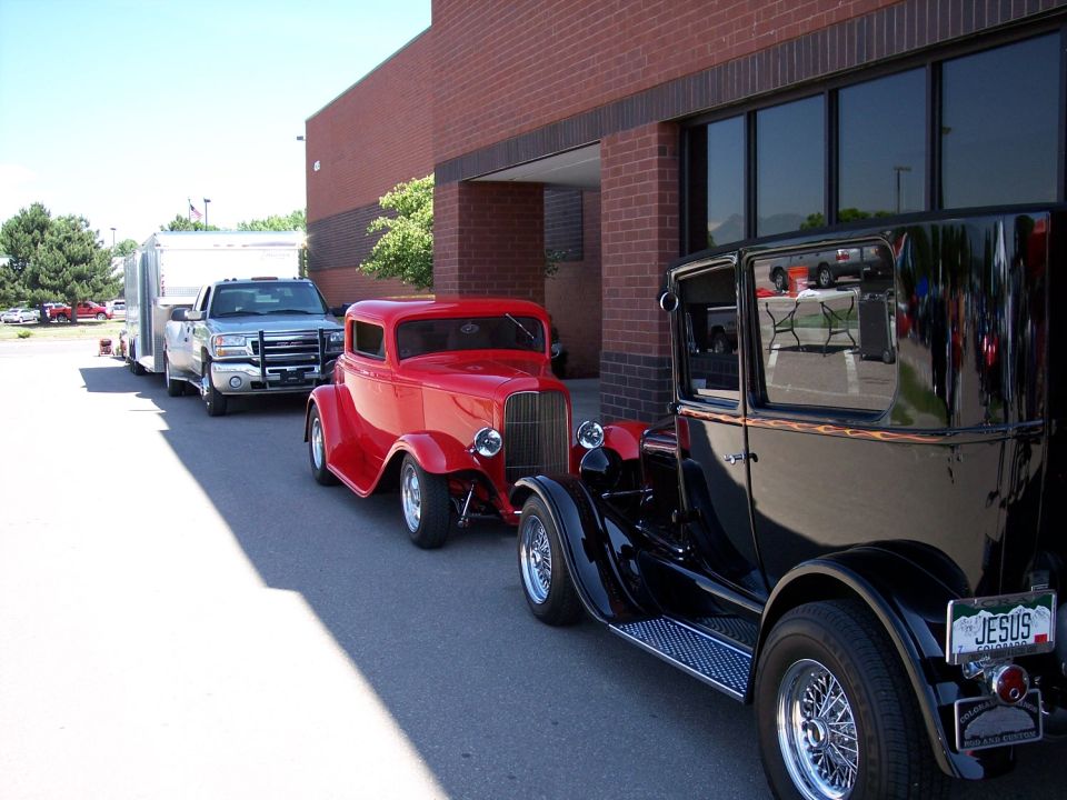 Silver pickup truck towing a trailer, red hot rod, and black vintage car parked in a line