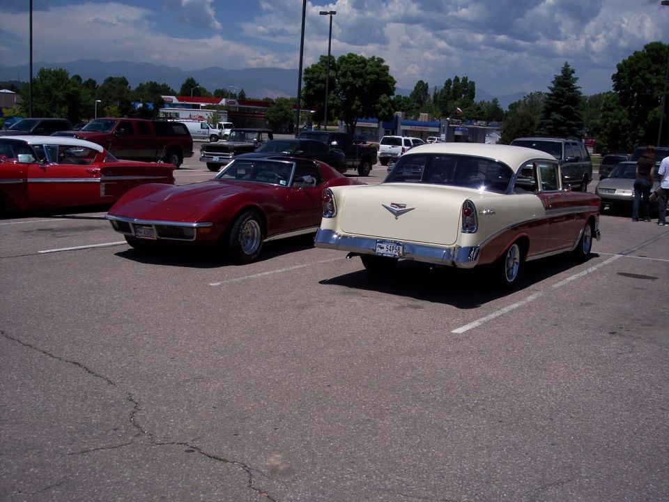 Two-tone beige and brown 1956 Chevrolet sedan parked, rear view. Red sports car to the left