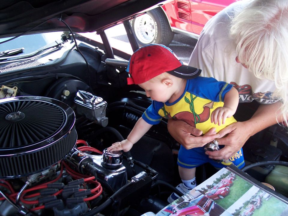 Baby in red cap and blue/yellow shirt reaches into car engine bay. Adult holds baby. Another car visible in background
