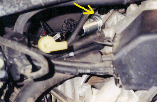 Engine bay close-up with yellow arrow pointing to a cylindrical metal sensor among wiring and hoses