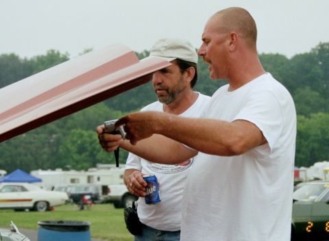 1976 Ford Ranchero - Two men are focused on a camera, with the hood of a vehicle open above them. Other cars and RVs are visible in the background.