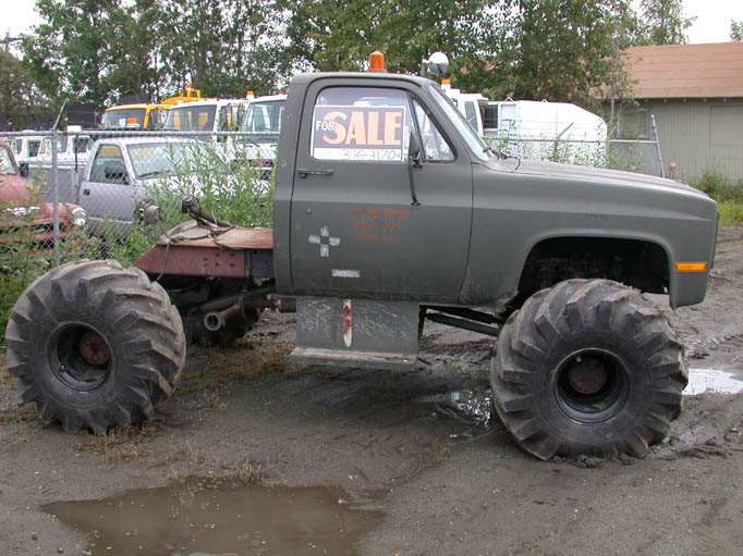 Olive-green military Chevy truck cab on giant tractor tires with a For Sale sign, bed removed