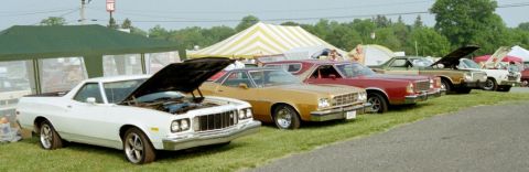 1976 Ford Ranchero - A line of classic cars, including a white 1976 Ford Ranchero, are parked on a grassy field under tents.