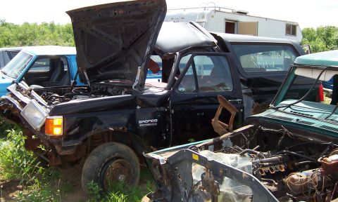 1980 Jeep CJ5 - This heavily damaged black Ford Bronco sits in a junkyard with its hood open, surrounded by other vehicles.