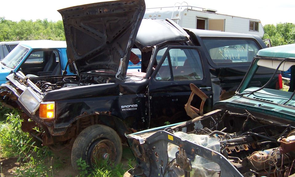 wrecked black Ford Bronco with hood up, parked next to other junked vehicles