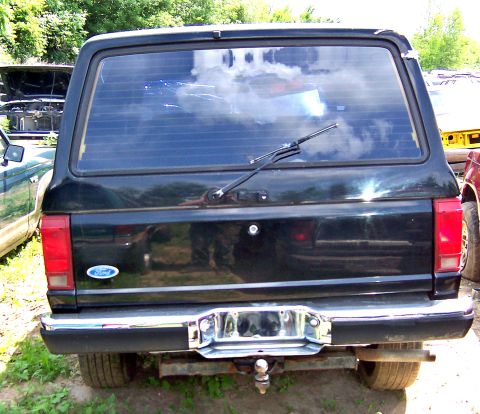 1980 Jeep CJ5 - The rear of a black Ford Bronco is visible, parked in a junkyard with other vehicles.