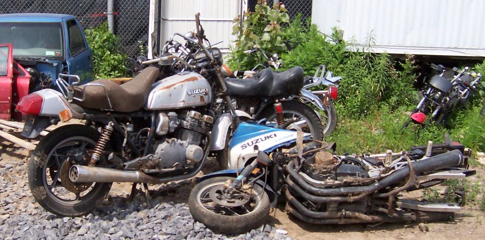 Several old Suzuki motorcycles are piled up in a junkyard. One is mostly intact, another is partially disassembled, and a third is visible in the background