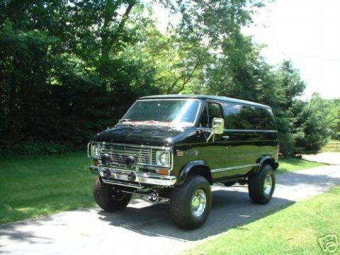 1978 Ford E250 - A black van with a lifted suspension and large off-road tires sits on a paved driveway surrounded by trees.
