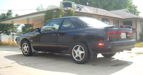 1989 Mazda MX-6 - A black 1989 Mazda MX-6 coupe is seen from the rear quarter angle in front of a house.
