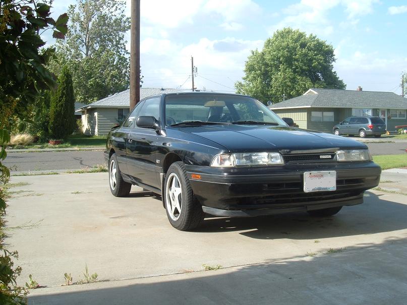 Black 1989 Mazda MX-6 coupe parked on a concrete driveway, front 3/4 view. The car has a license plate and a GT badge