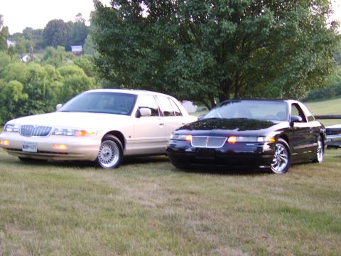 1996 Mercury Grand Marquis - Two large sedans, a Mercury Grand Marquis and a Lincoln, are parked on a grassy lawn under a large tree.