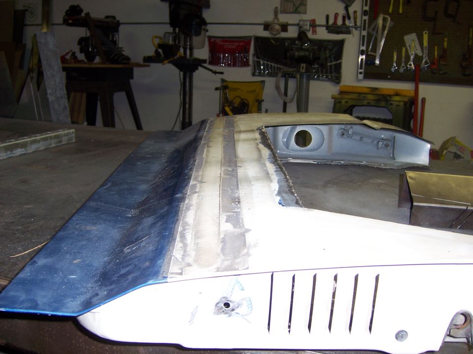 Car hood with blue paint and body filler work, next to a white fender with vertical vents. Garage setting with tools on wall