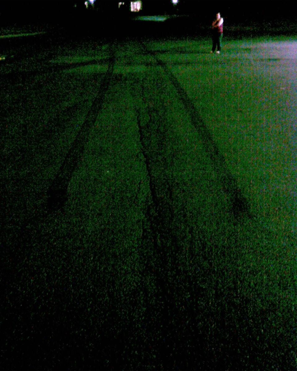 Tire skid marks across dark grass at night with a distant figure