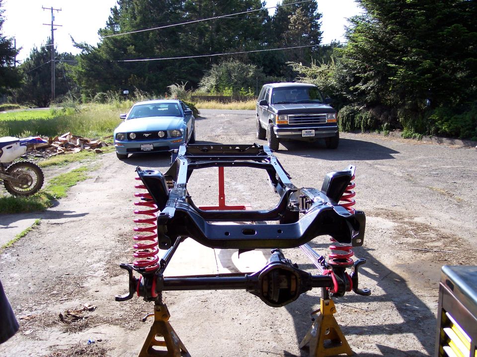 Vehicle frame on jack stands with a blue car and a dark SUV in the background
