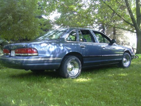 1996 Ford Crown Victoria - A blue sedan sits on a grassy lawn under trees, viewed from the rear passenger side.