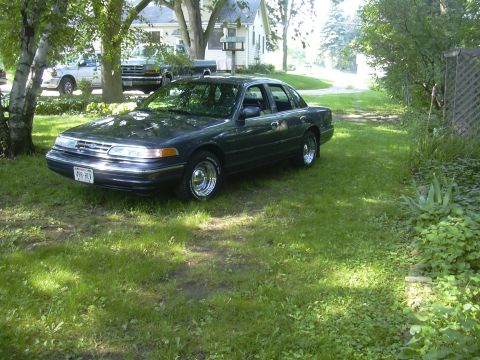 1996 Ford Crown Victoria - Dark sedan parked on a grassy area next to a house, with a white pickup truck visible behind it.