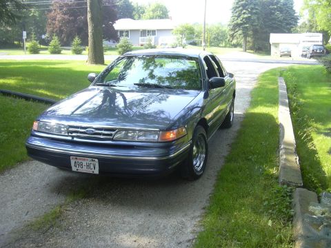 1996 Ford Crown Victoria - A blue sedan is parked on a gravel driveway next to a grassy lawn and a concrete curb.