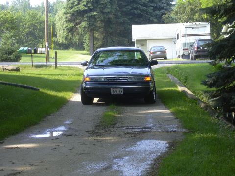 1996 Ford Crown Victoria - A dark blue sedan is parked on a gravel driveway, with other vehicles and a house visible in the background.