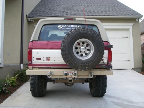 1995 Ford Bronco - Rear view of a lifted red and tan Ford Bronco in a driveway, wearing a tan rear tube bumper with D-ring shackles and a swing-out carrier ho…