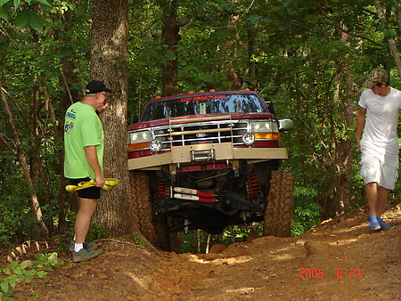 1995 Ford Bronco - A lifted red truck navigates a steep, muddy off-road trail with two onlookers.