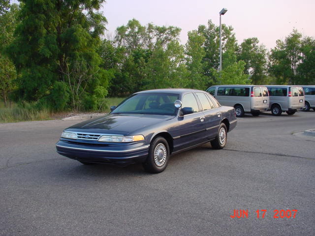 Dark blue 2003 Ford Crown Victoria police car parked on asphalt, with silver vans in the background