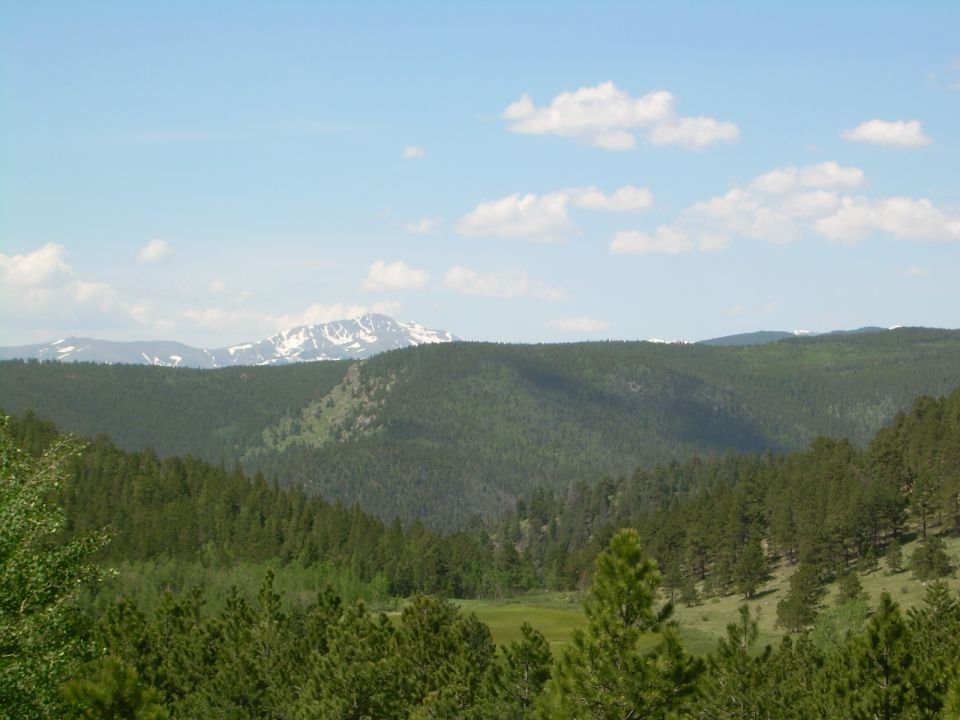 Mountain landscape with snow-capped peaks and pine forests under a blue sky with clouds. Text overlay reads SUPERMOTORS