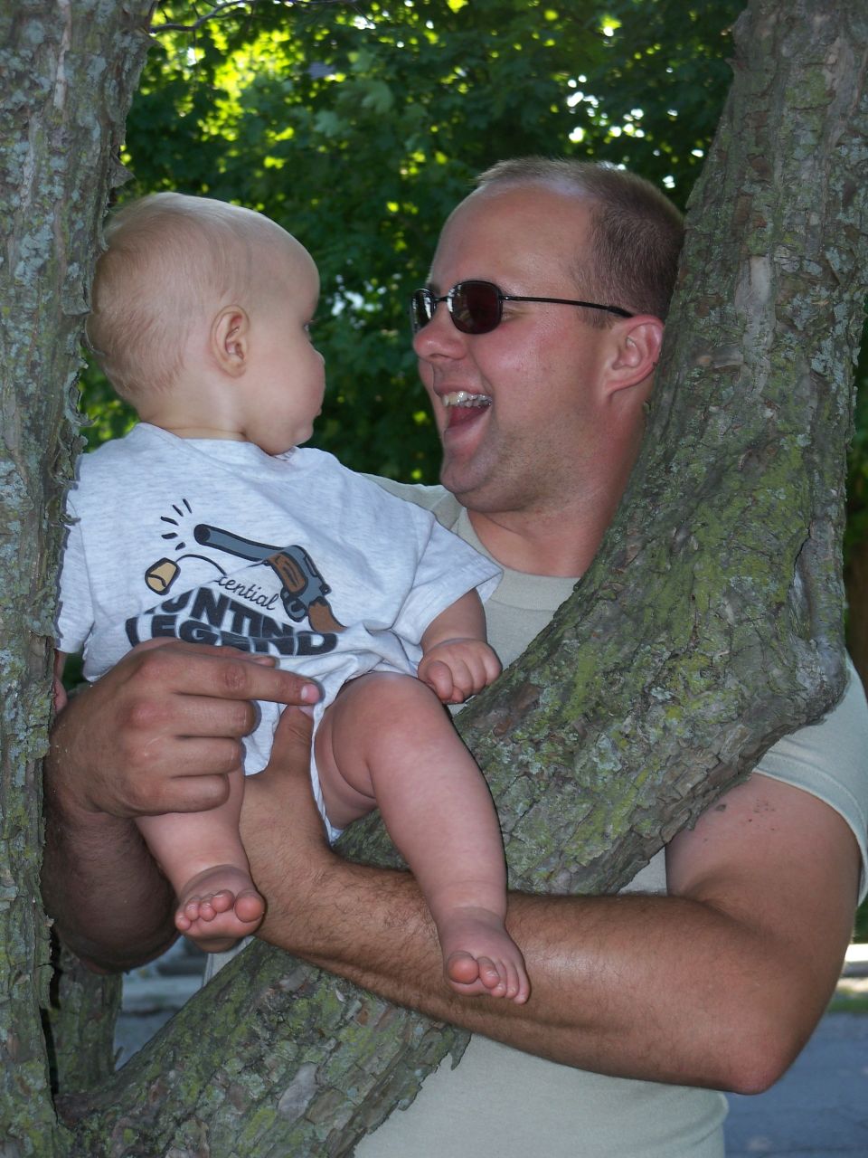 Man holding baby in a tree, baby wears shirt with gun graphic
