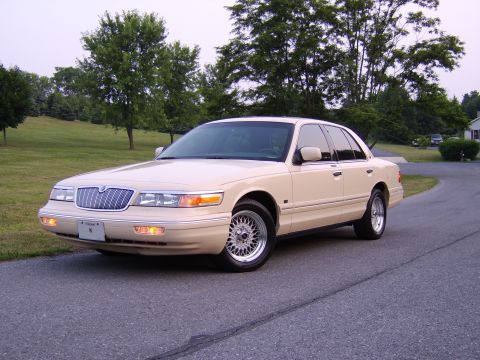 1996 Mercury Grand Marquis - Cream 1996 Mercury Grand Marquis sedan parked on a paved road with a grassy hill and trees behind it.