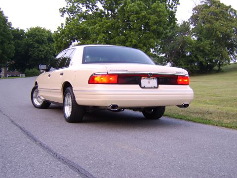 1996 Mercury Grand Marquis - Rear view of a tan sedan with chrome wheels and dual exhaust pipes, parked on a road with trees in the background.