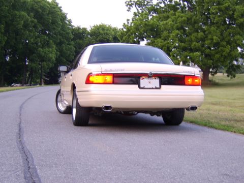 1996 Mercury Grand Marquis - The rear of a sedan is visible, parked on a paved road with a grassy area and trees behind it.