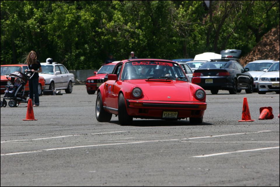 Red Porsche 911 with racing banner on windshield, driving on a track with cones. Other cars and a woman with a stroller are in the background