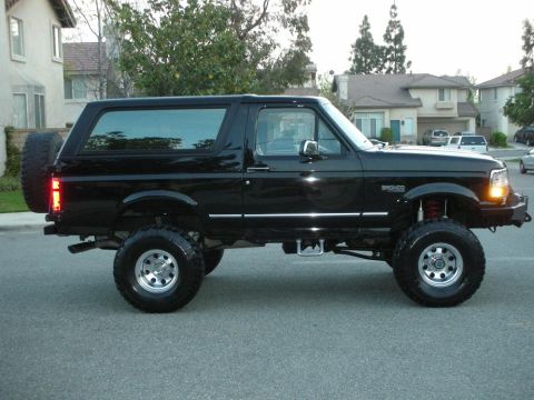 1996 Ford Bronco - This lifted black Ford Bronco is shown from the side, parked on a suburban street with houses in the background.