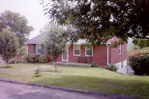 1991 Ford LTD Crown Victoria - A suburban brick house is seen on a cloudy day, surrounded by trees and a well-kept lawn.