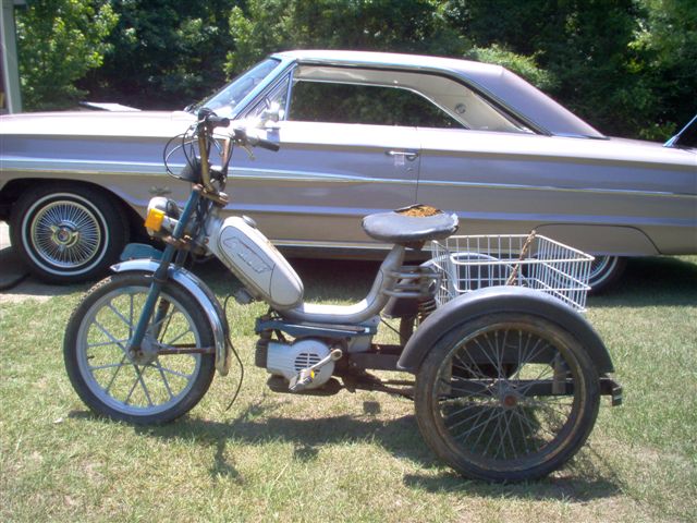 Three-wheeled moped with a basket and a light purple 1964 Ford Galaxie 500 behind it
