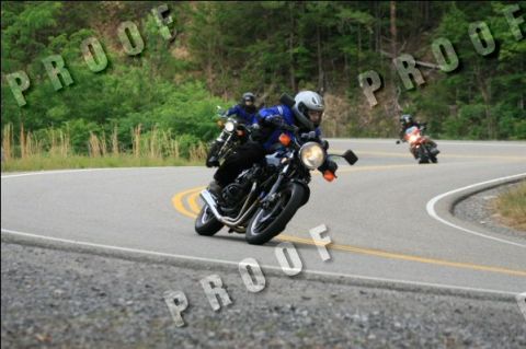 1985 Ford LTD Crown Victoria - Several motorcycles navigate a sharp turn on a scenic, wooded highway.