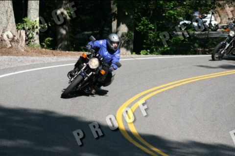 1985 Ford LTD Crown Victoria - Motorcyclist navigates a sharp turn on a paved road surrounded by trees.