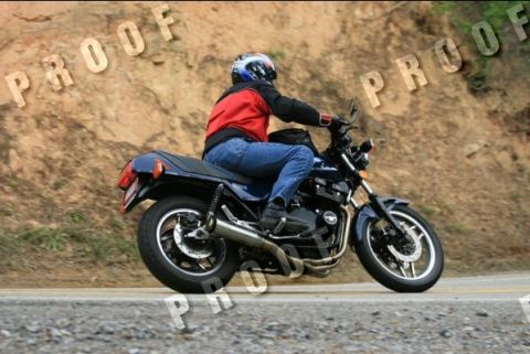 1985 Ford LTD Crown Victoria - A rider on a dark blue motorcycle cruises down a paved road with a rocky hillside in the background.