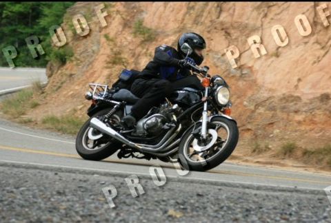 1985 Ford LTD Crown Victoria - A rider on a black motorcycle navigates a curve on a road bordered by a rocky embankment.