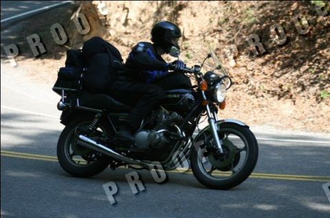 1985 Ford LTD Crown Victoria - Rider on a motorcycle with luggage bags on the rear rack travels down a road.