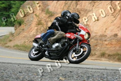 1985 Ford LTD Crown Victoria - Two people ride a red motorcycle on a paved road next to a rocky hillside.