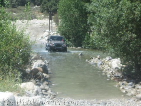 1987 Suzuki Samurai - A Jeep Wrangler navigates a stream on a sunny day, surrounded by trees and rocky terrain.