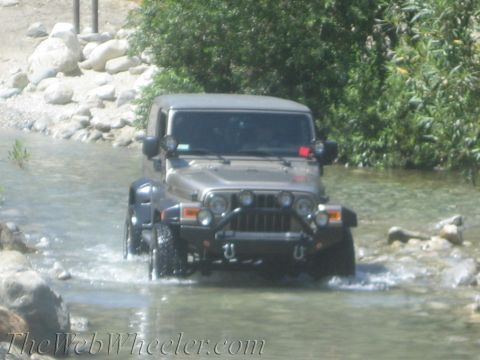 1987 Suzuki Samurai - A tan Jeep Wrangler navigates a rocky stream, its front grille and bumper clearly visible.