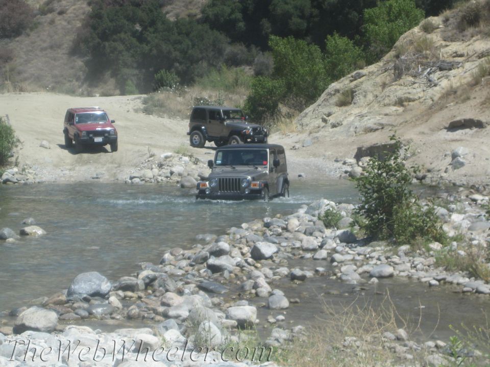 Three Jeeps driving through a rocky stream bed on a dirt path