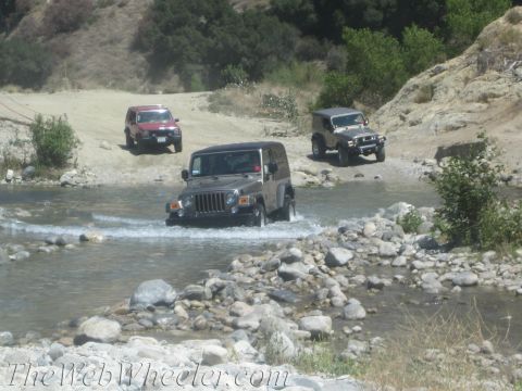 1987 Suzuki Samurai - Several Jeep Wranglers navigate a shallow river on a sunny day, with rocky terrain and sparse vegetation surrounding them.