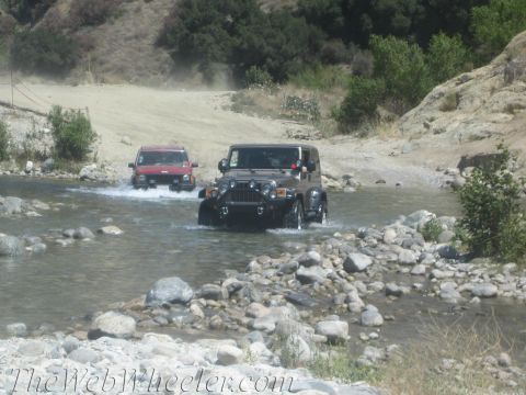 1987 Suzuki Samurai - Two off-road vehicles navigate a shallow, rocky stream in a dry, hilly landscape.