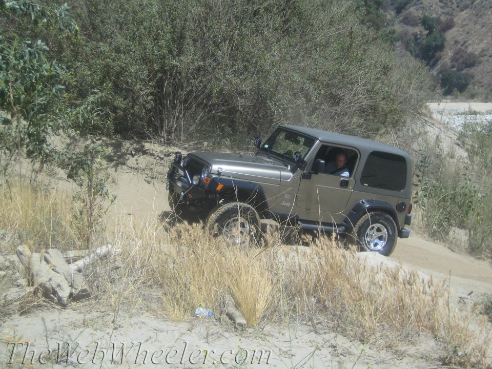 Jeep Wrangler driving on a sandy, off-road trail with dry brush and hills in the background