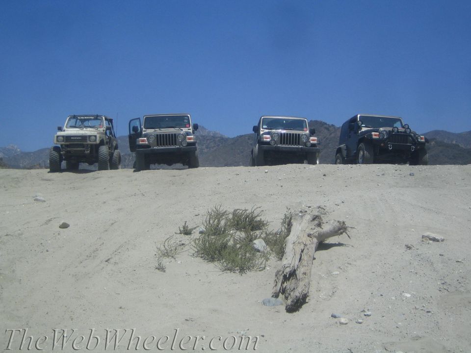 Four off-road vehicles parked on a sandy hill under a clear blue sky. One is a white Suzuki Samurai, the others are Jeeps
