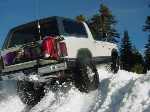 1986 Ford Bronco - A white 1980s Ford Bronco navigates a snowy terrain, equipped with snowboards and a spare gas can.