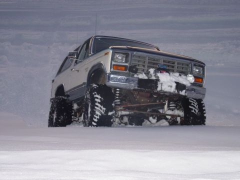 1986 Ford Bronco - Lifted white and blue Ford Bronco climbing a snowbank at dusk, shot low from the passenger 3/4 front so the coil springs, radius arms and t…