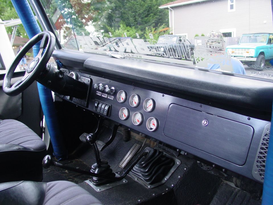 Interior of a 1973 Ford Bronco showing dashboard, steering wheel, gauges, and gear shifter. Two other Broncos are visible outside